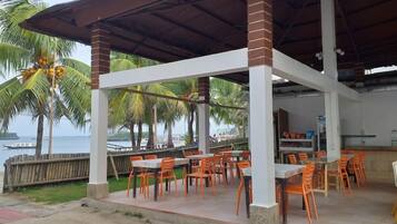 An outdoor seating area with tables and chairs, a covered structure, and a view of the sea and palm trees.