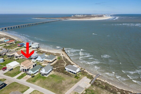 An aerial view of a coastal residential area with houses, a beach, and a long pier extending into the ocean.