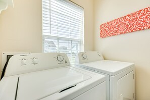 A laundry area with two white washing machines, a window with blinds, and a red patterned wall decoration.