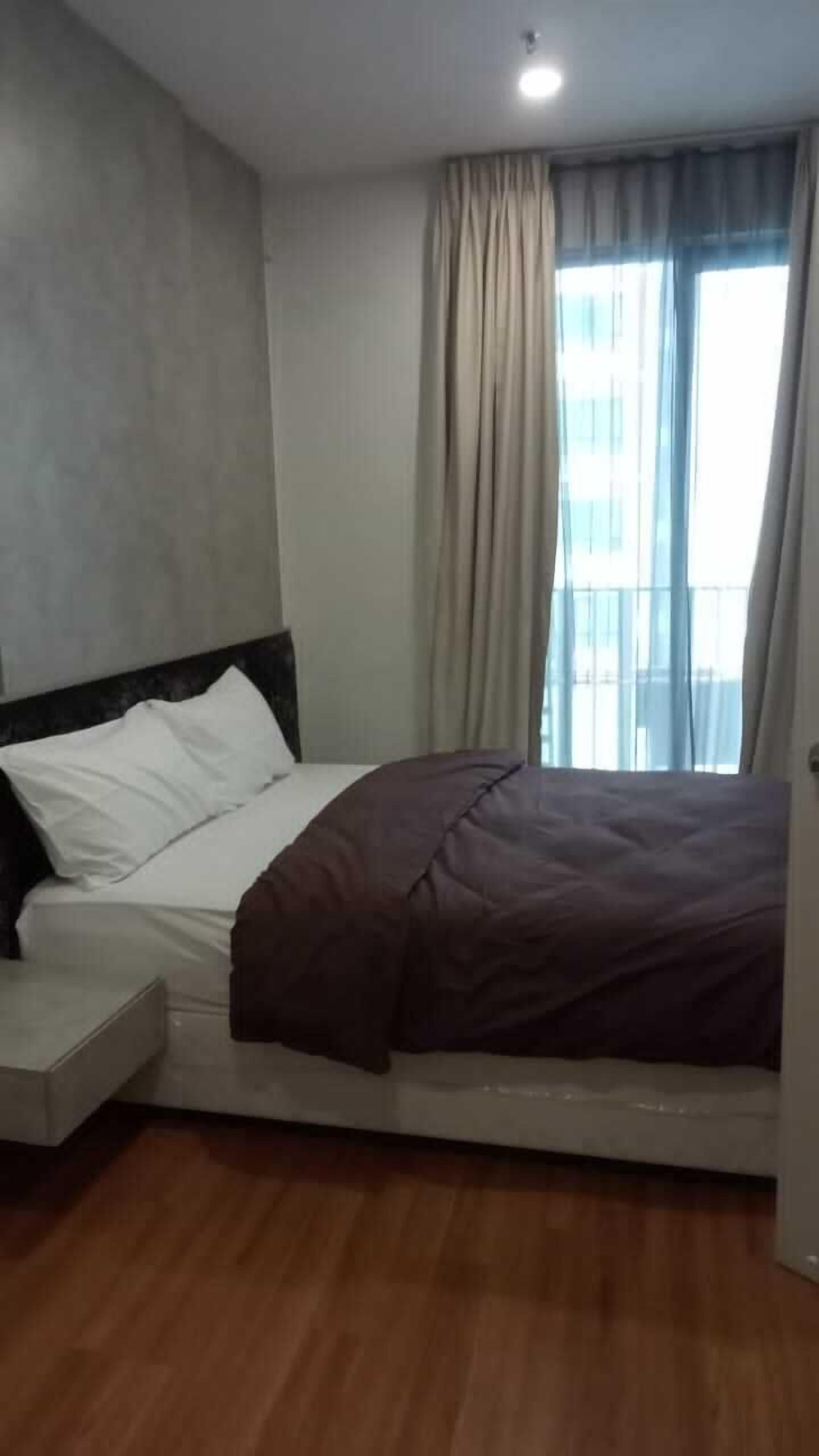 Una habitación de hotel con cama, mesita de noche, ventana con cortinas y vistas a edificios.