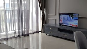 Un saló modern amb un televisor de pantalla plana sobre una consola gris, finestres grans amb cortines lleugeres i un sòl net revestit de rajoles.