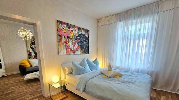 Un dormitorio con una cama, una mesita de noche con lámpara, un cuadro en la pared y una ventana con cortinas.