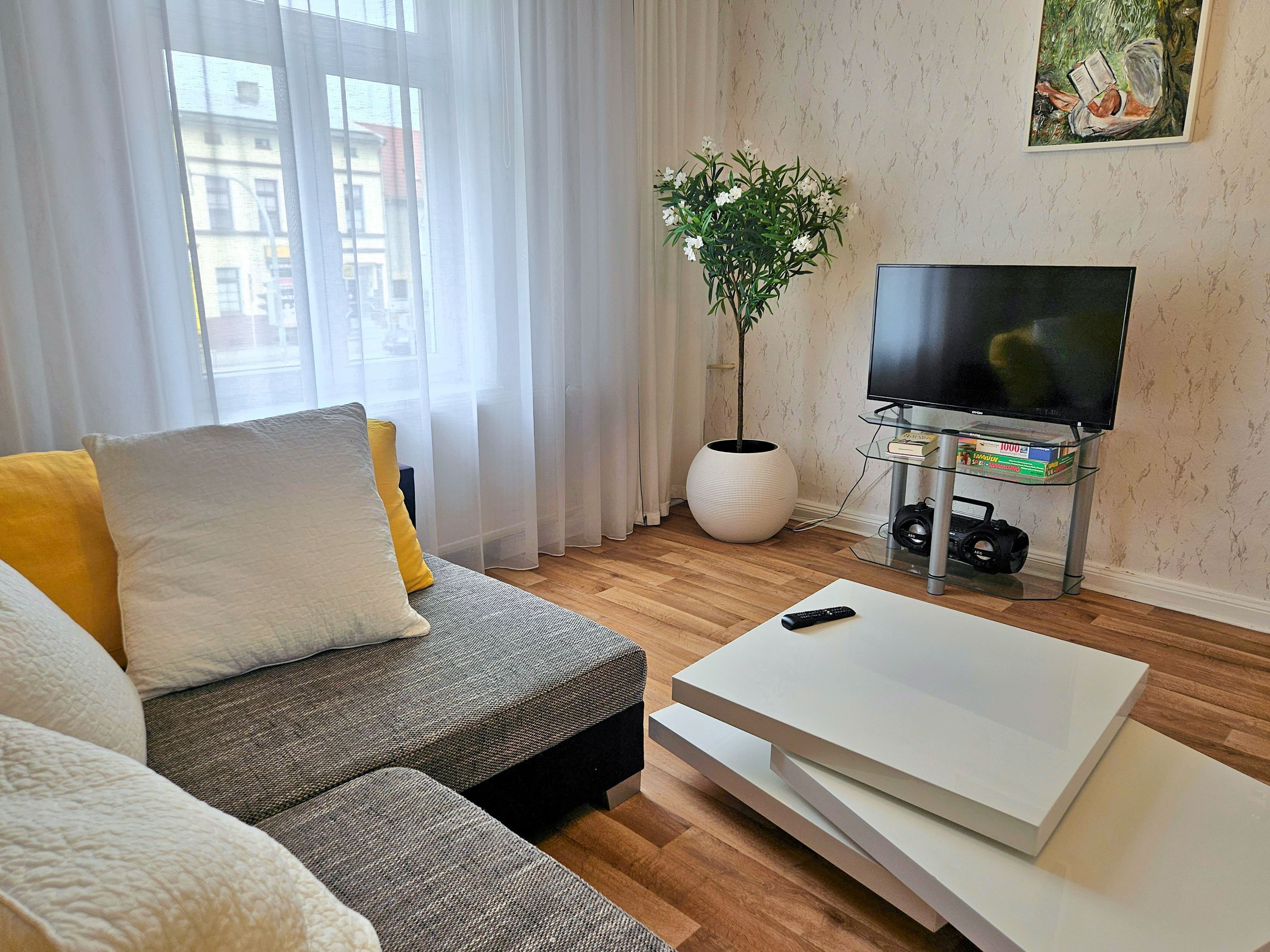Una sala de estar con un sofá, una mesa de centro, un televisor sobre un mueble, una planta y un cuadro en la pared.