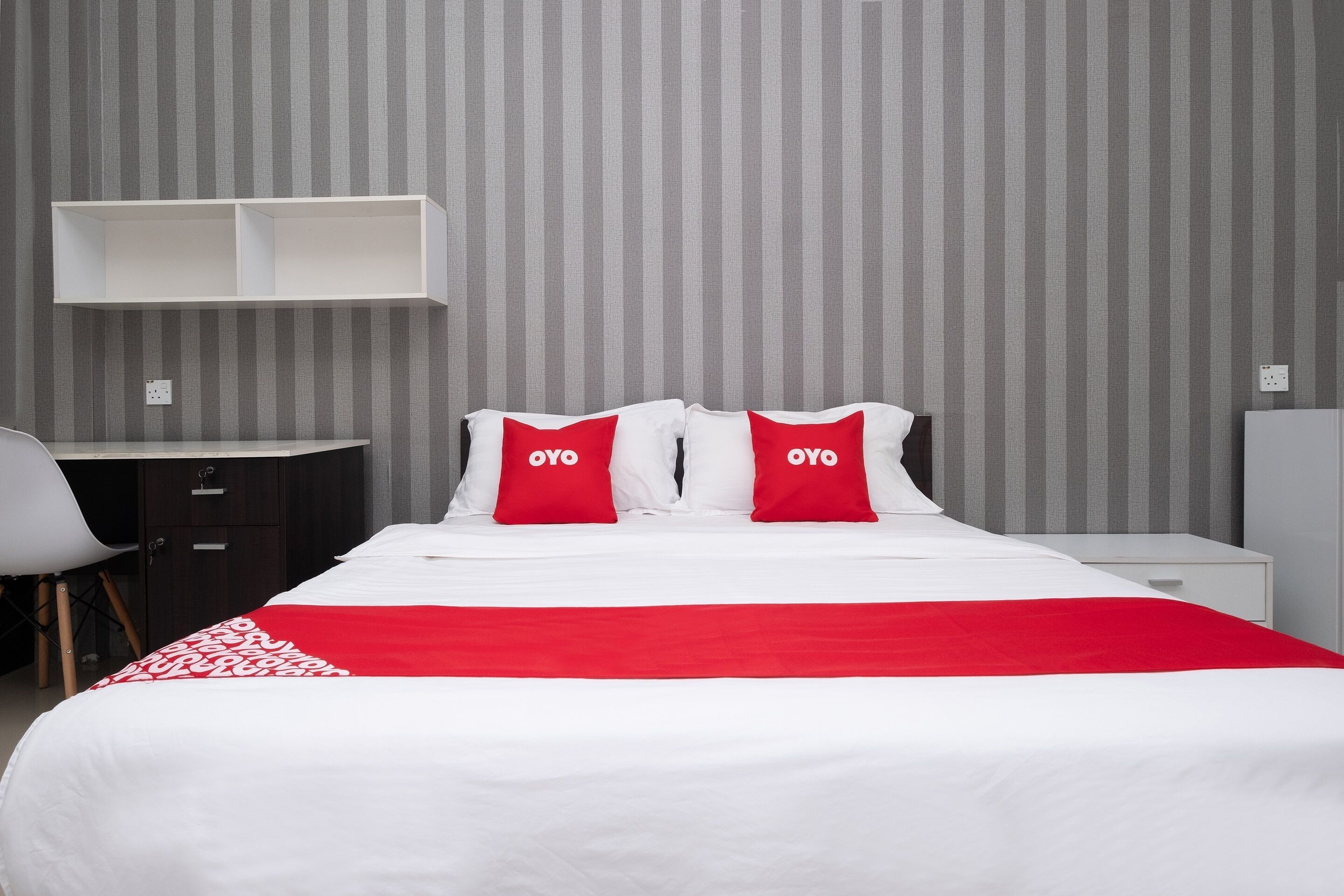 A hotel room with a bed, two red pillows, a white bedspread, a desk with a chair, and a wardrobe.
