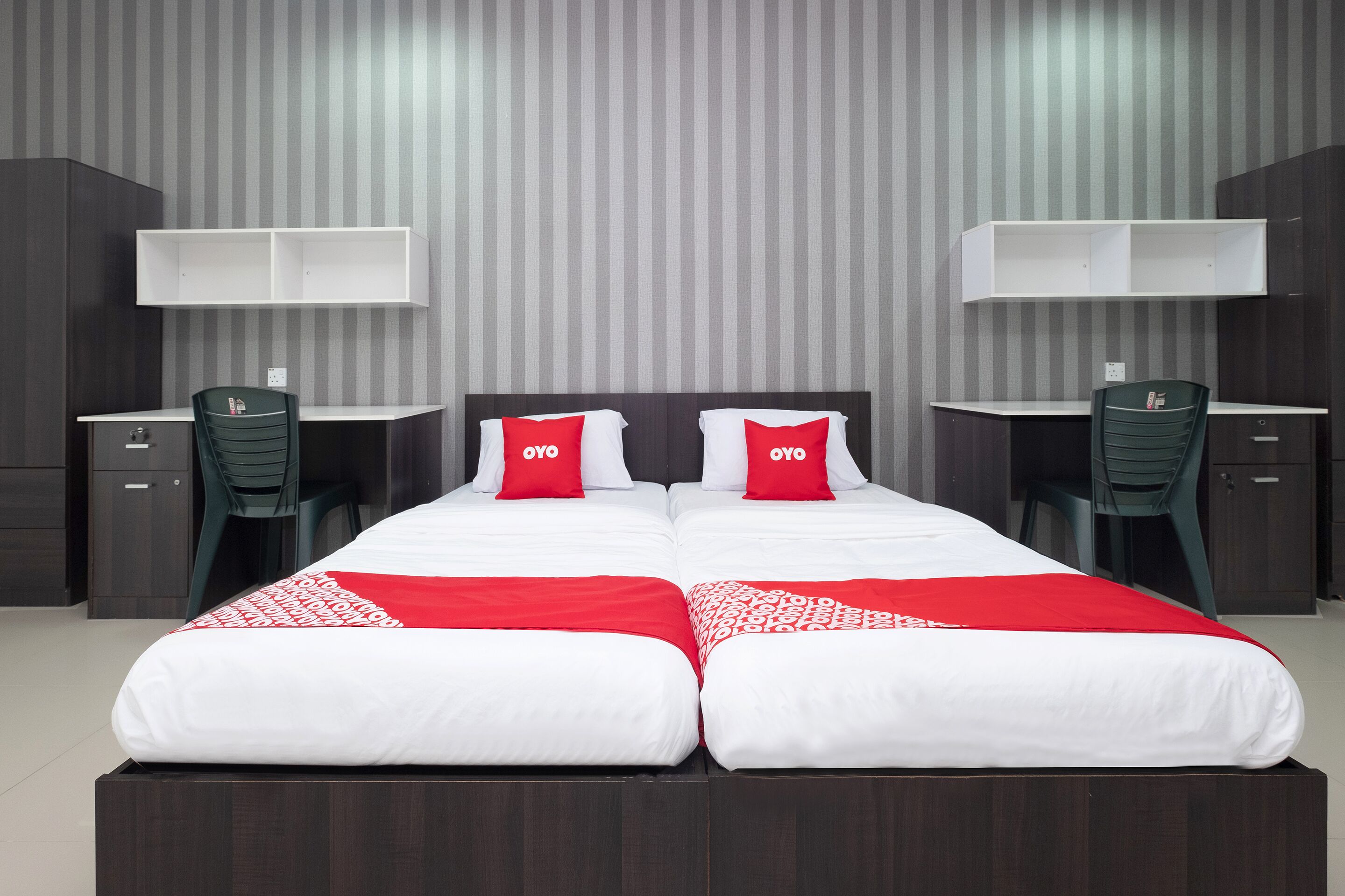 Un lit double avec deux oreillers rouges, un couvre-lit blanc et une couverture rouge. Il y a deux bureaux avec des chaises, une armoire et une étagère fixée au mur.