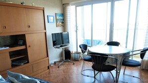 Ein Zimmer mit einem Bett, einem Fernseher auf einem Schrank, einem Esstisch mit Stühlen und einem Fenster mit Vorhängen.