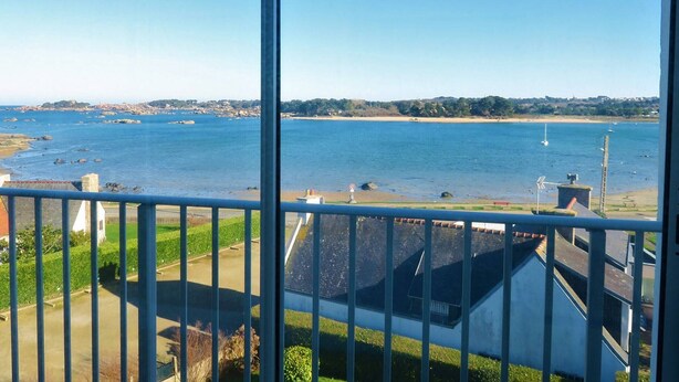 Blick aufs Meer von einem Balkon mit Geländer. Man sieht ruhiges Wasser, einen Sandstrand und Gebäude in der Ferne.