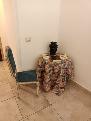 Quarto de canto com uma cadeira, uma mesinha com toalha florida e uma estátua.