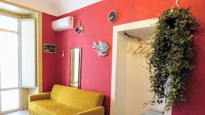Una stanza con un divano giallo, un letto con copripiumino decorato, un piccolo armadietto grigio e una pianta fissata al muro.