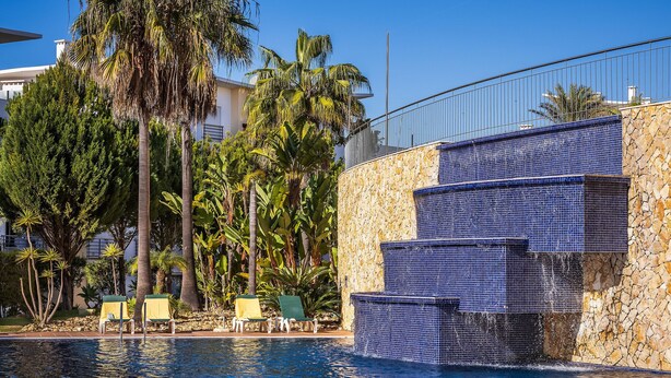A swimming pool with a stone wall and blue mosaic tiles, surrounded by palm trees and lounge chairs. - Flat Amora OCV - Ocean View (Albufeira)