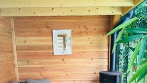 A cozy wooden cabin with a sofa, cushions, a woven ottoman, and a wood-burning stove.