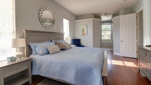 Quarto com cama grande, ventoinha no teto, mesa de cabeceira com luminária, cômoda e porta para outro espaço.