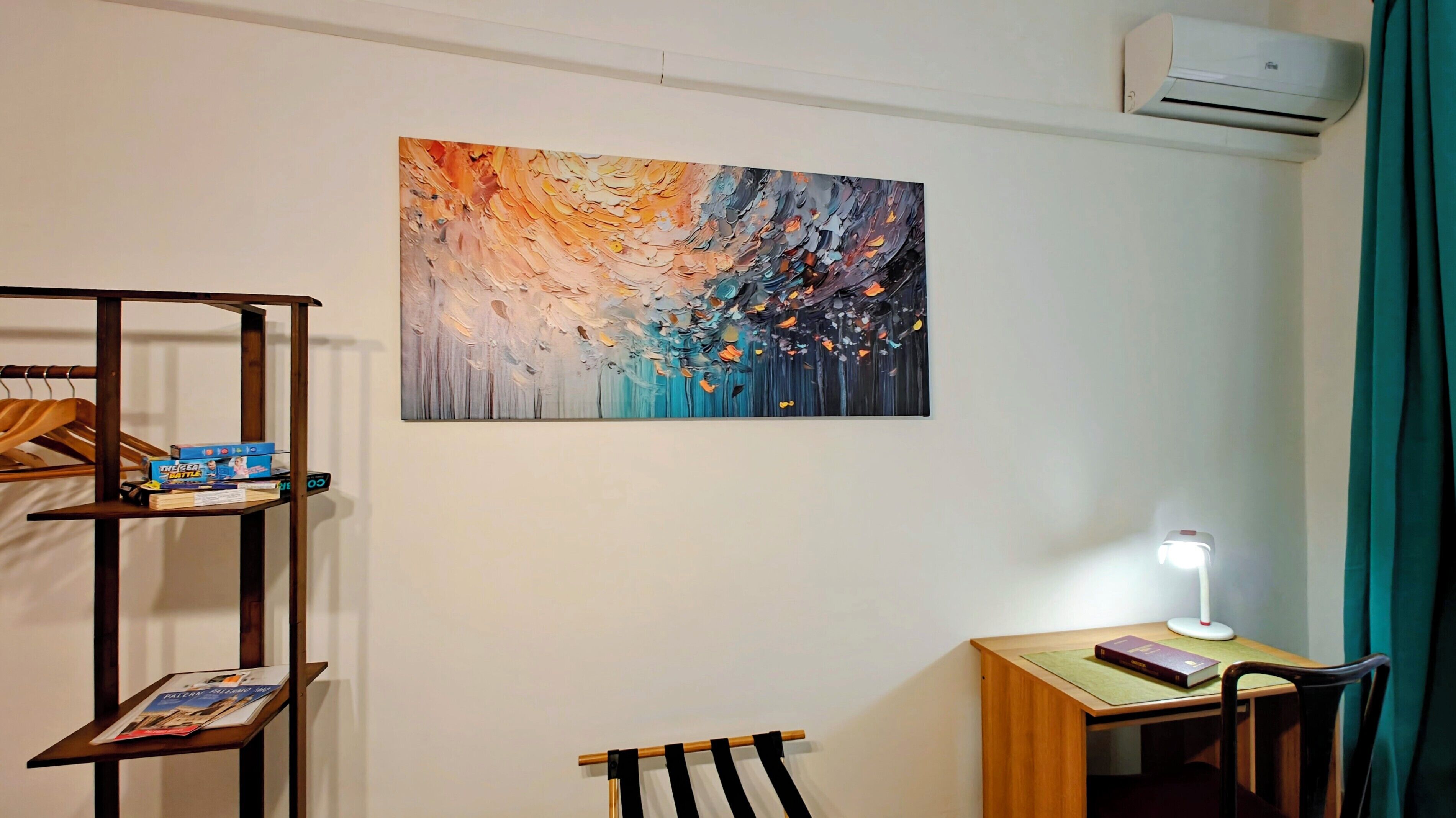 A room with a painting, a desk with a lamp, a chair, and a hanging shelf with items.