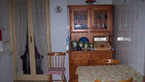 Un comedor con una mesa cubierta con mantel floreado, dos sillas de madera, un mueble de madera con puertas de vidrio y una silla con asiento a cuadros.