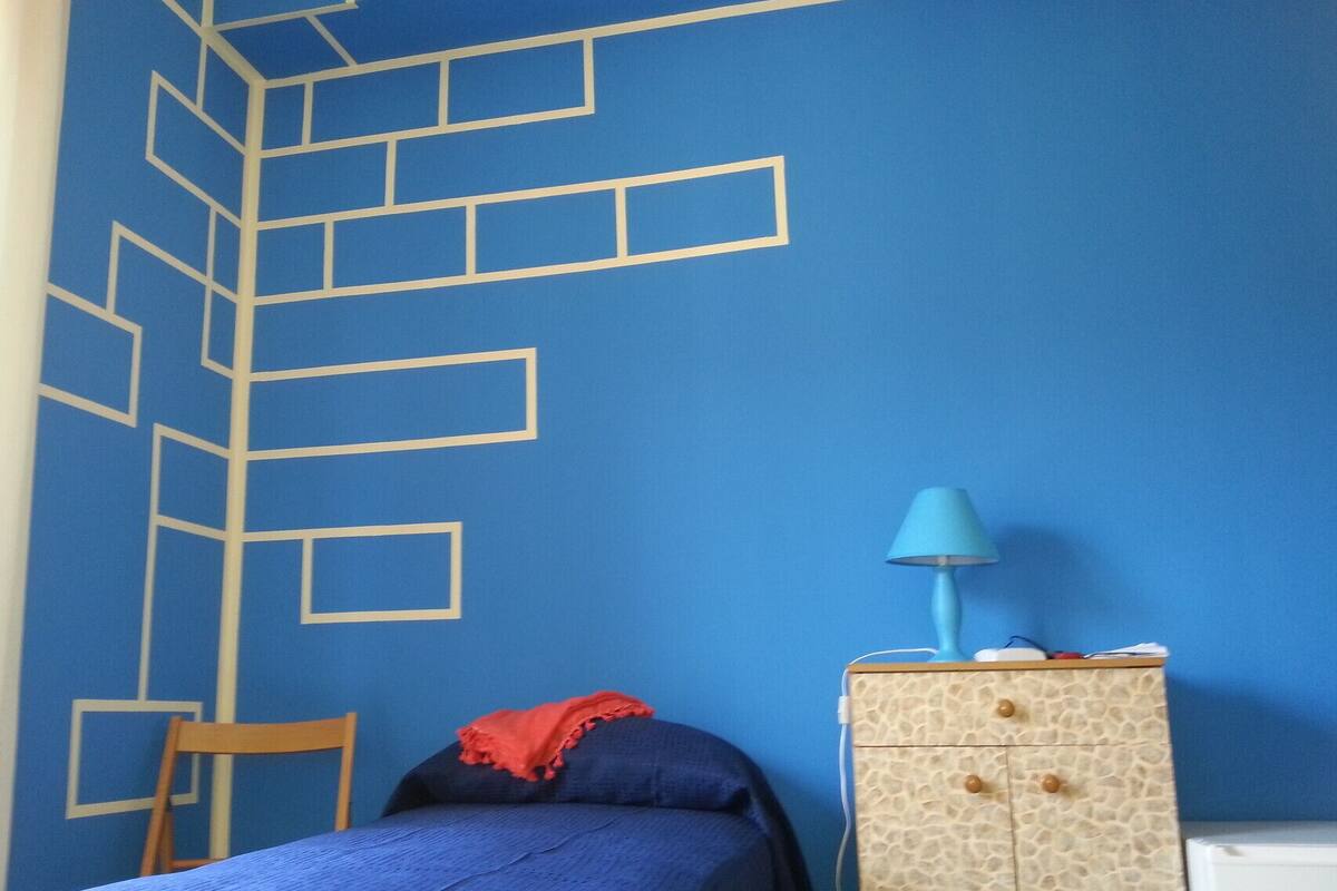 A bedroom with a blue wall, a bed, a chair, a dresser, and a lamp.
