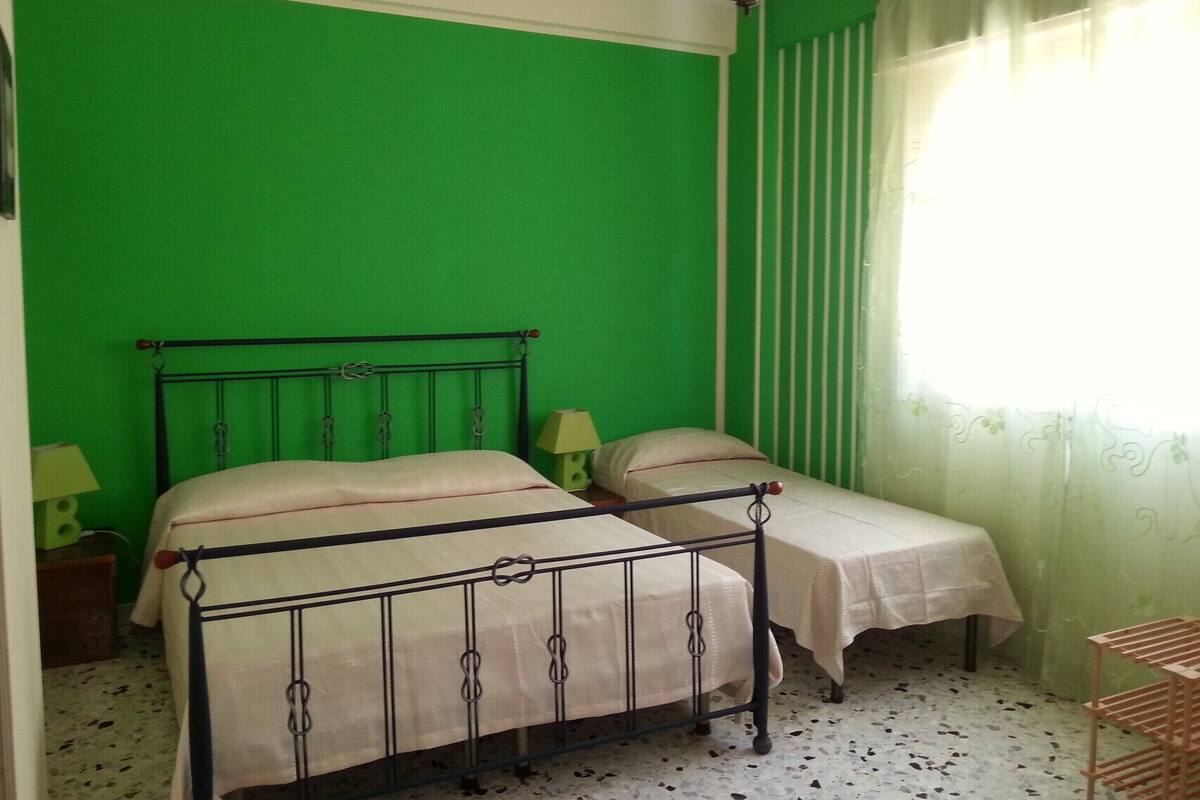 A room with a green wall, two beds with white linens, a chandelier, and a window with sheer curtains.