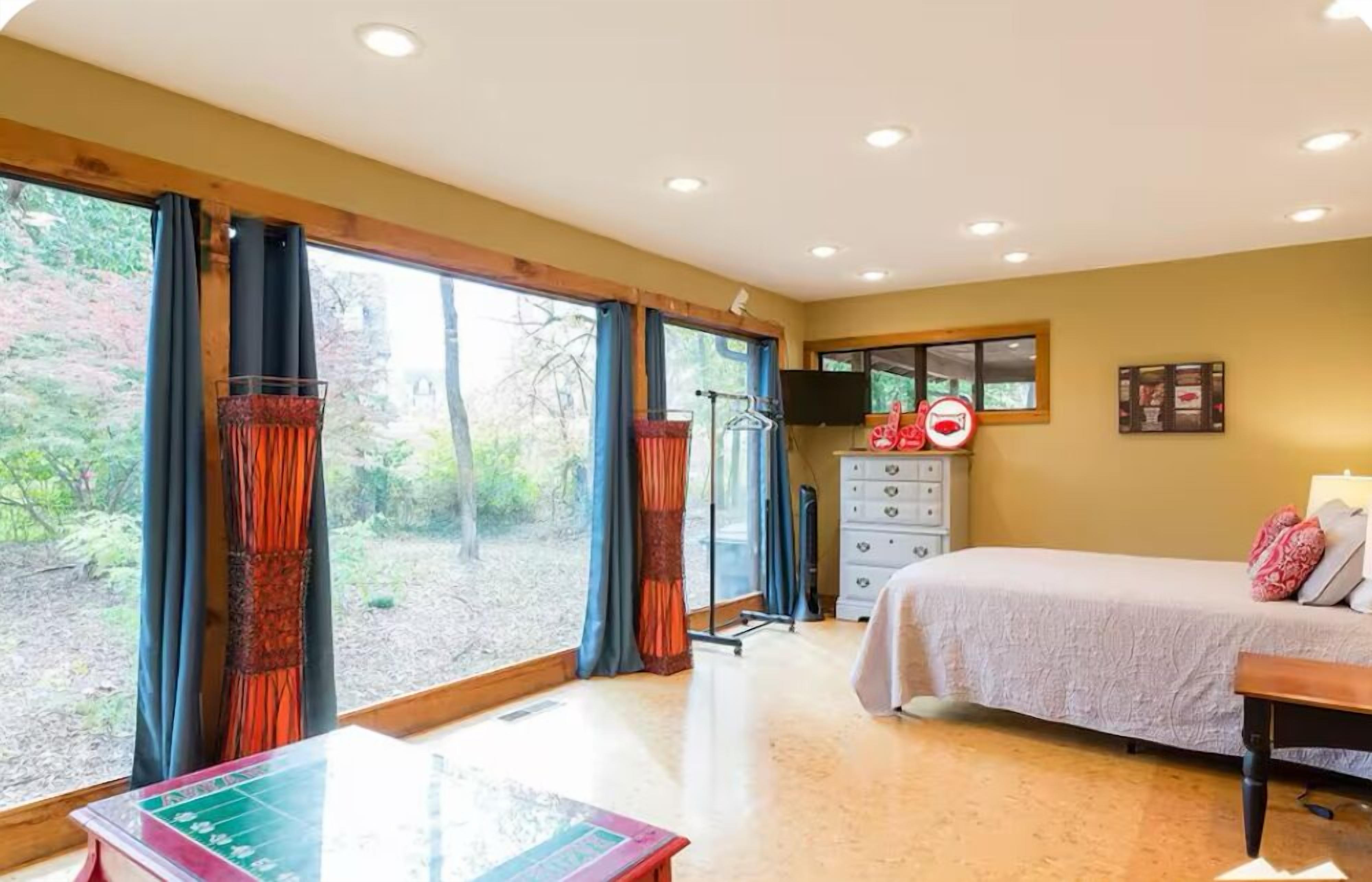 A bedroom with a bed, dresser, and a desk with a chair. There are large windows with curtains, a TV on the wall, and a no-smoking sign.