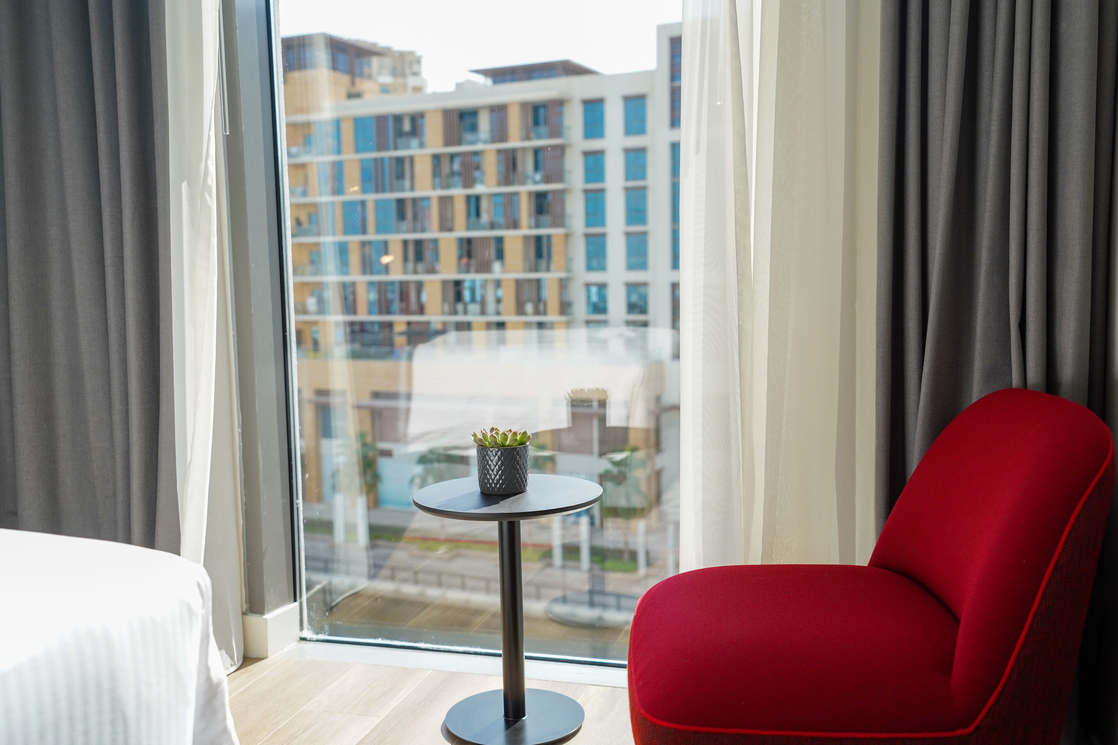 Una habitación de hotel moderna con un ventanal grande, una silla roja, una mesita redonda con una planta y una cama.