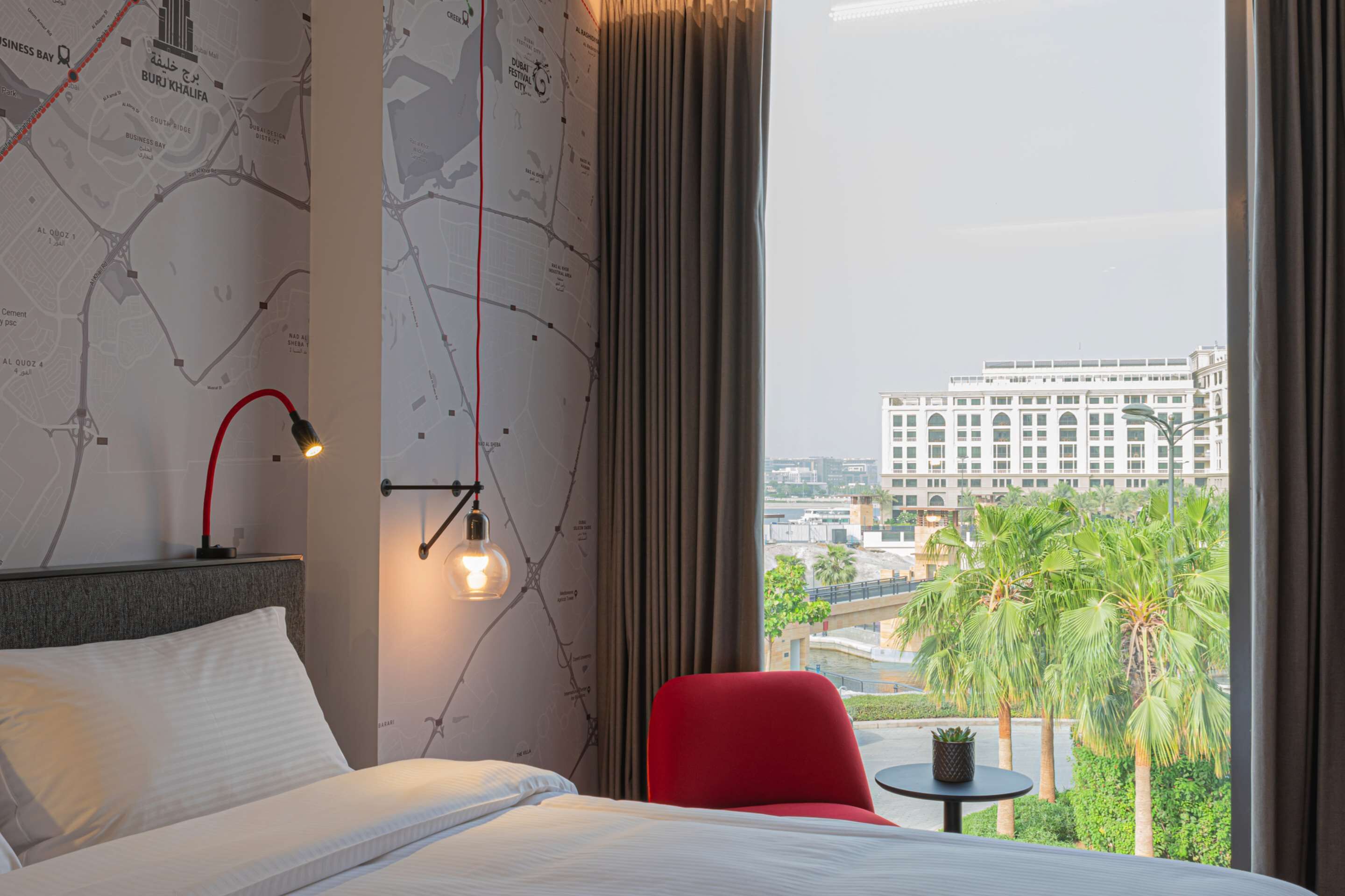 A hotel room with a bed, a red chair, a small table, a map on the wall, and a view of a building and palm trees outside.