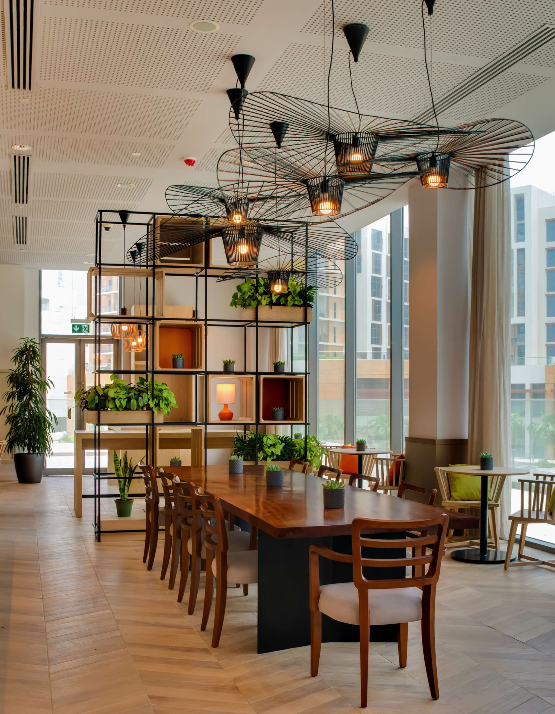 A modern dining area with a long wooden table, black metal shelving with plants, and pendant lighting.