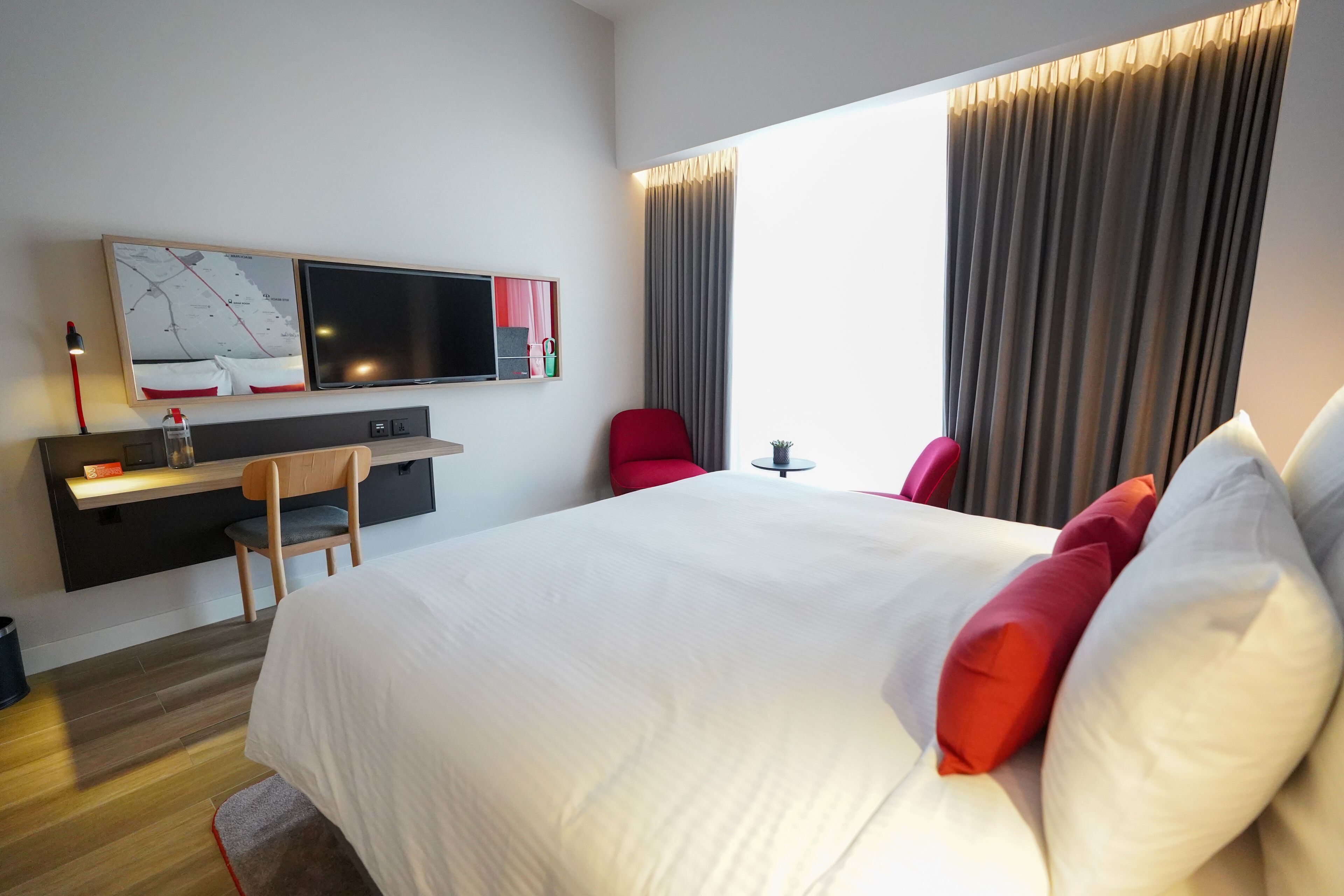 A modern hotel room with a large bed, a desk with a chair, a TV, and a window with curtains.