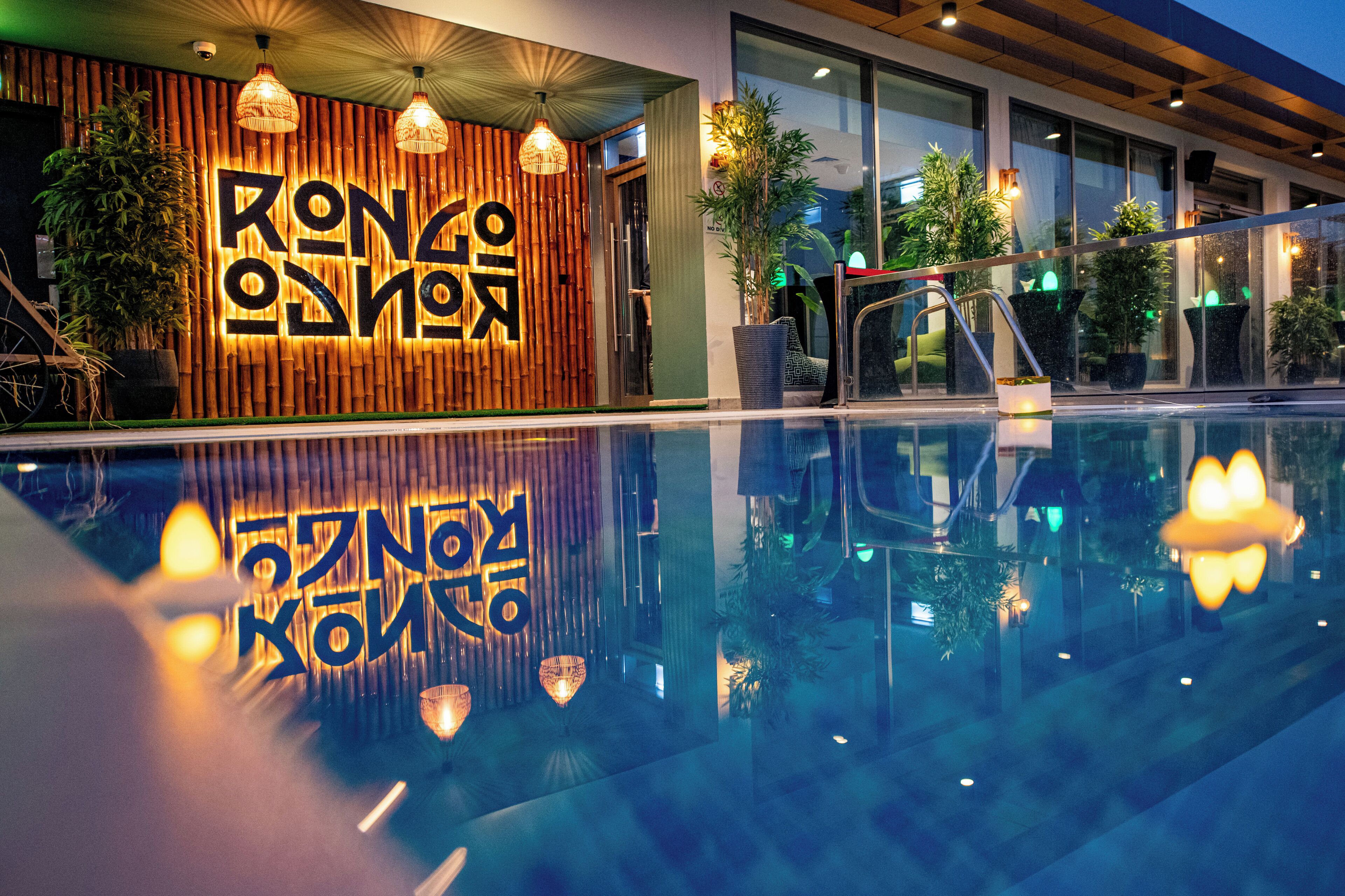 Entrada moderna de hotel com parede de bambu e letreiro iluminado, piscina refletindo as luzes e plantas em vasos ao redor.