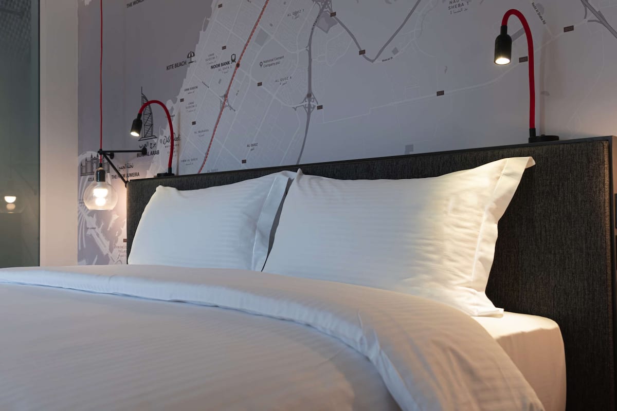 Un lit bien fait, avec du linge de lit blanc, une tête de lit dotée d’un panneau foncé et une carte murale avec une ligne rouge.