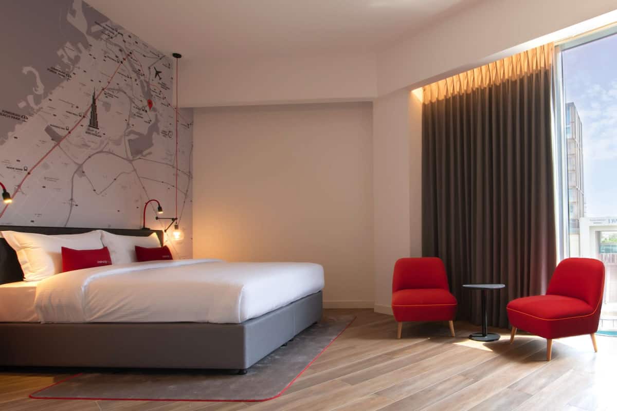 Une chambre d’hôtel moderne avec un grand lit, une carte accrochée au mur, deux fauteuils rouges et une vue sur la ville par la fenêtre.