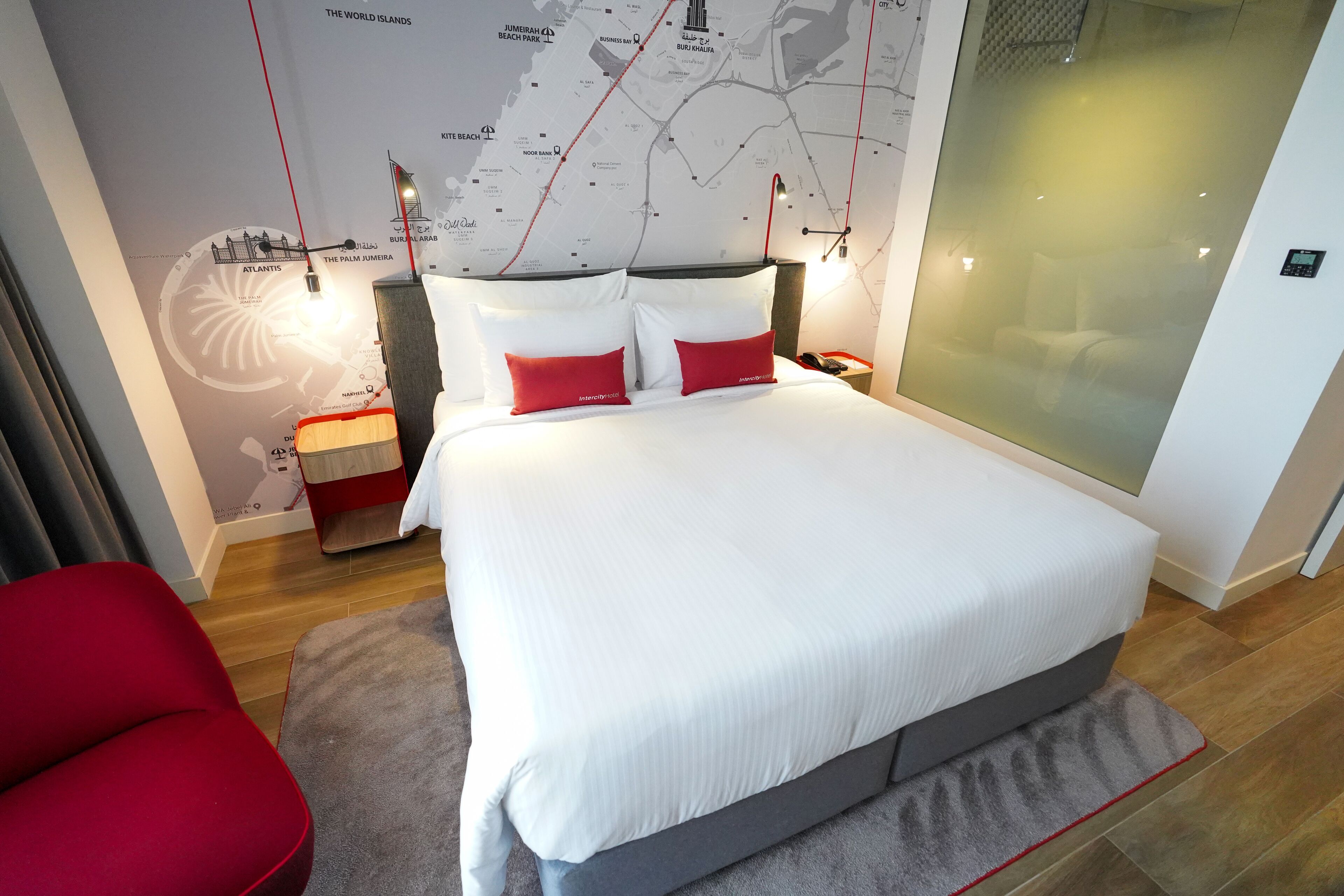 A modern hotel room with a large bed, red and white pillows, a red chair, a map on the wall, and a nightstand with a lamp.