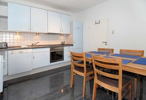 A modern kitchen with white cabinets, a built-in oven, and a dining area with wooden chairs and a table. - Holiday house North Sea magic (Esens)
