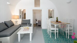 A modern living room with a sofa, coffee table, and dining table. There is a lamp, a chair, and a small plant on the table. - Cave Agapi house with private hot tub and view of the Oia caldera (Oia)