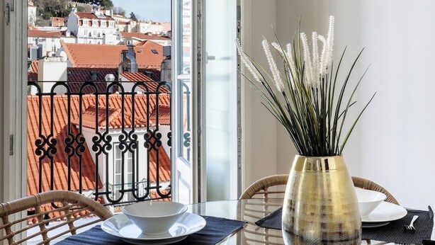 En matplats med ett bord dukat för två personer, en vas med vita blommor och en utsikt över byggnader med röda tak genom ett fönster.