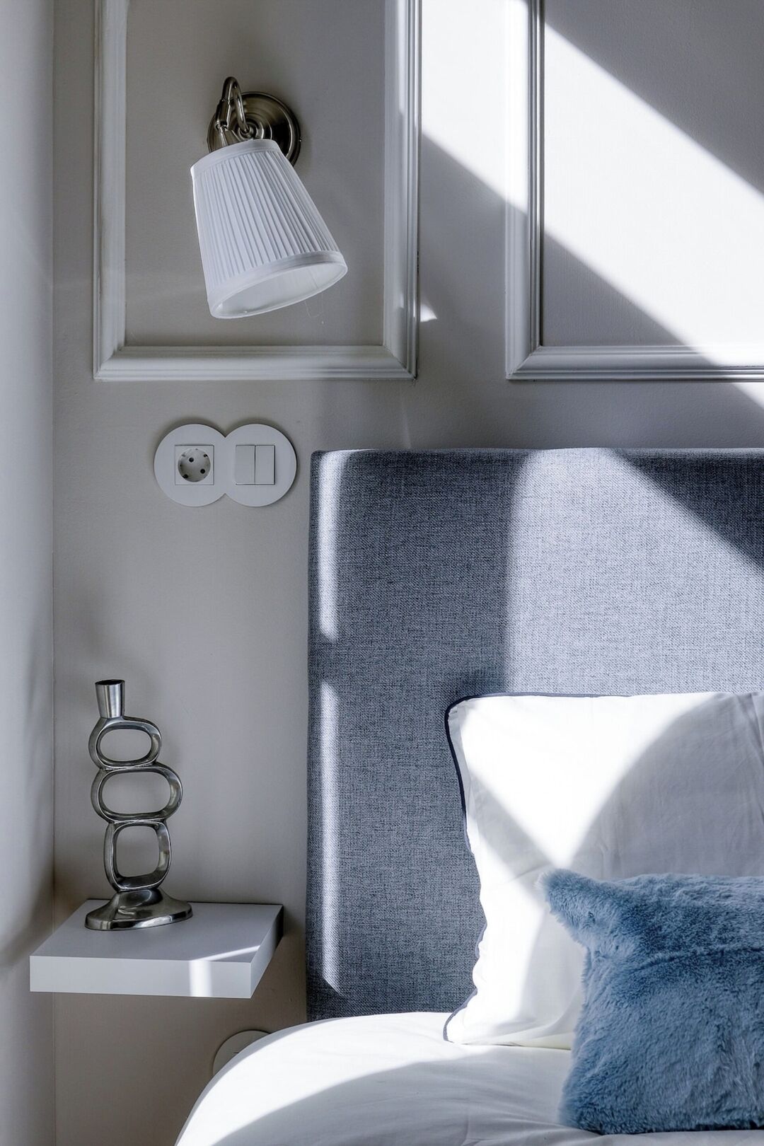 Un dormitorio con cama, lámpara de noche, enchufe y un objeto decorativo de metal sobre una mesita de noche.
