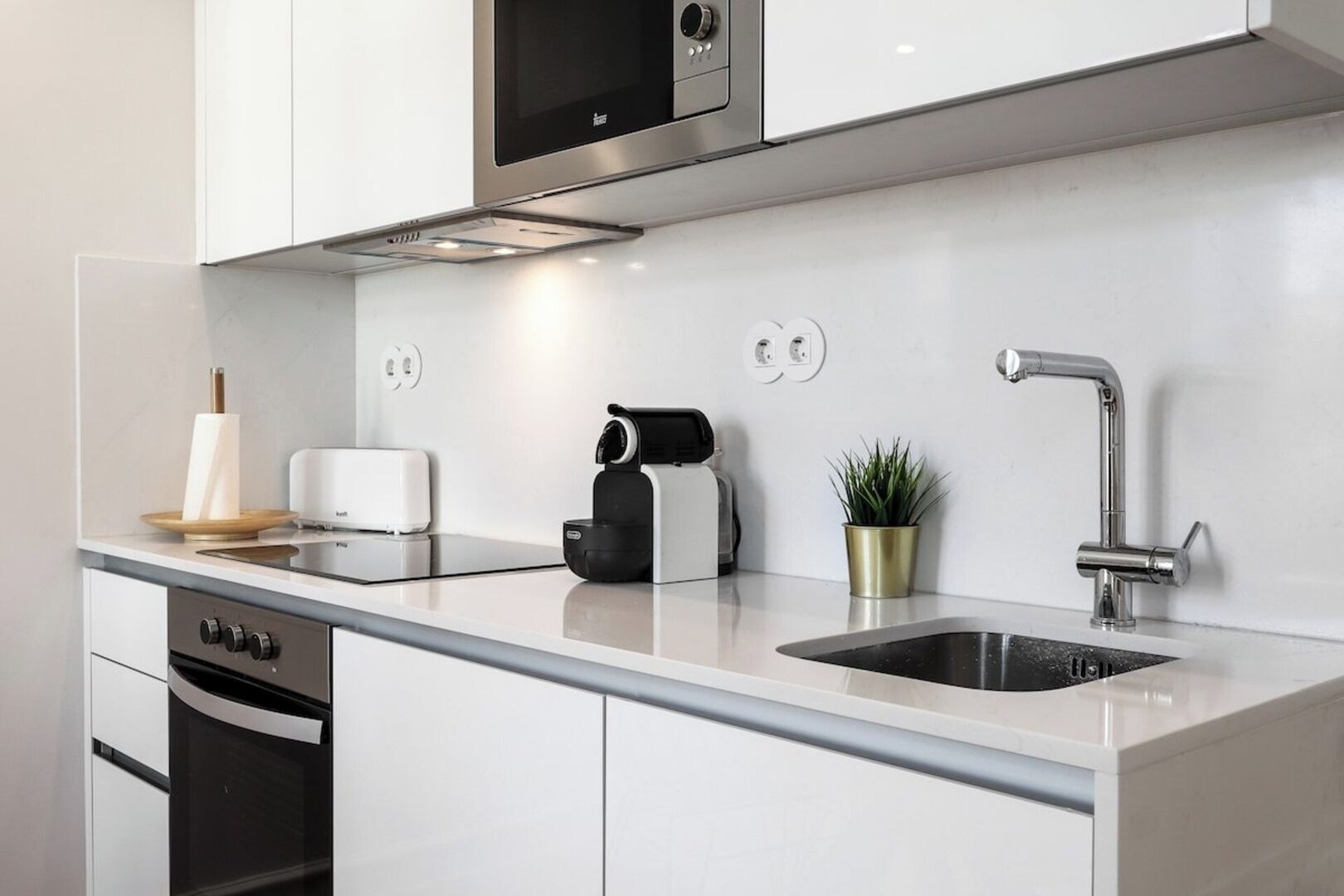 A modern kitchen with white cabinetry, a stainless steel range hood, a built-in toaster, a coffee machine, a sink with a modern faucet, and a potted plant.