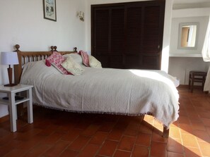 A bed with a white bedspread, two pillows, and a wooden headboard. There is a nightstand with a lamp and a small shelf. The room has a tiled floor and a large window with a view of the outside. - Villa Casa Monte, Quinta Do Paraiso, Carvoeiro Fibre WiFi (Carvoeiro)