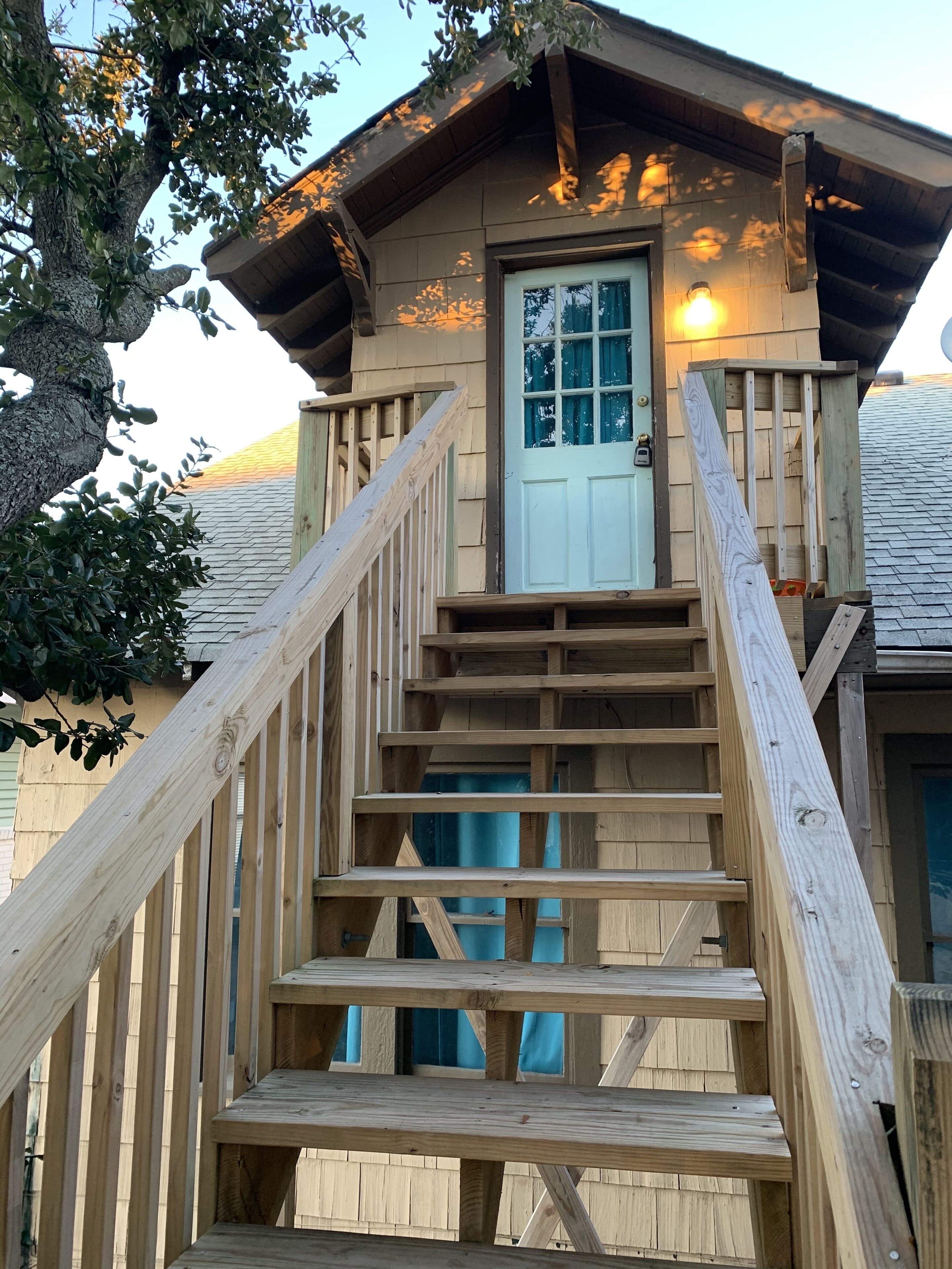 Un escalier en bois mène à une cabane dotée d’une porte et de fenêtres, le tout étant entouré d’arbres.