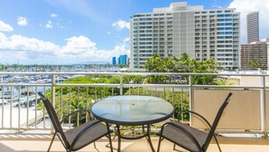 A balcony with a round glass table and two chairs, overlooking a marina with boats and a cityscape in the background. - Ilikai Hotel 1 Bedroom Yacht Harbor View, minutes to the Beach! (Honolulu)