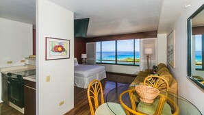 A compact hotel room with a kitchenette, a bed, a dining area with a glass table, and a large window offering a view of the ocean. - Discovery Bay Ocean View Studio, Near the Beach! (Honolulu)
