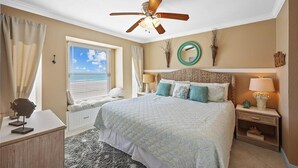 A bedroom with a bed, a ceiling fan, a window with curtains, a bedside table with a lamp, and a nightstand. - Seascape 1111 2 Bedroom Condo by RedAwning (Galveston)