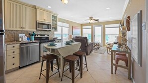 A modern kitchen with a central island, bar stools, and a dining area with a table and chairs. - Seascape 1111 2 Bedroom Condo by RedAwning (Galveston)