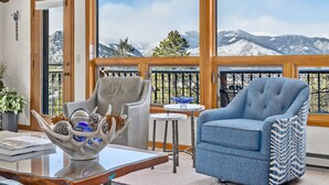 A room with a view of snow-covered mountains, a grey armchair, a blue armchair, a glass coffee table with decorative items, and a small round table with a blue glass object.