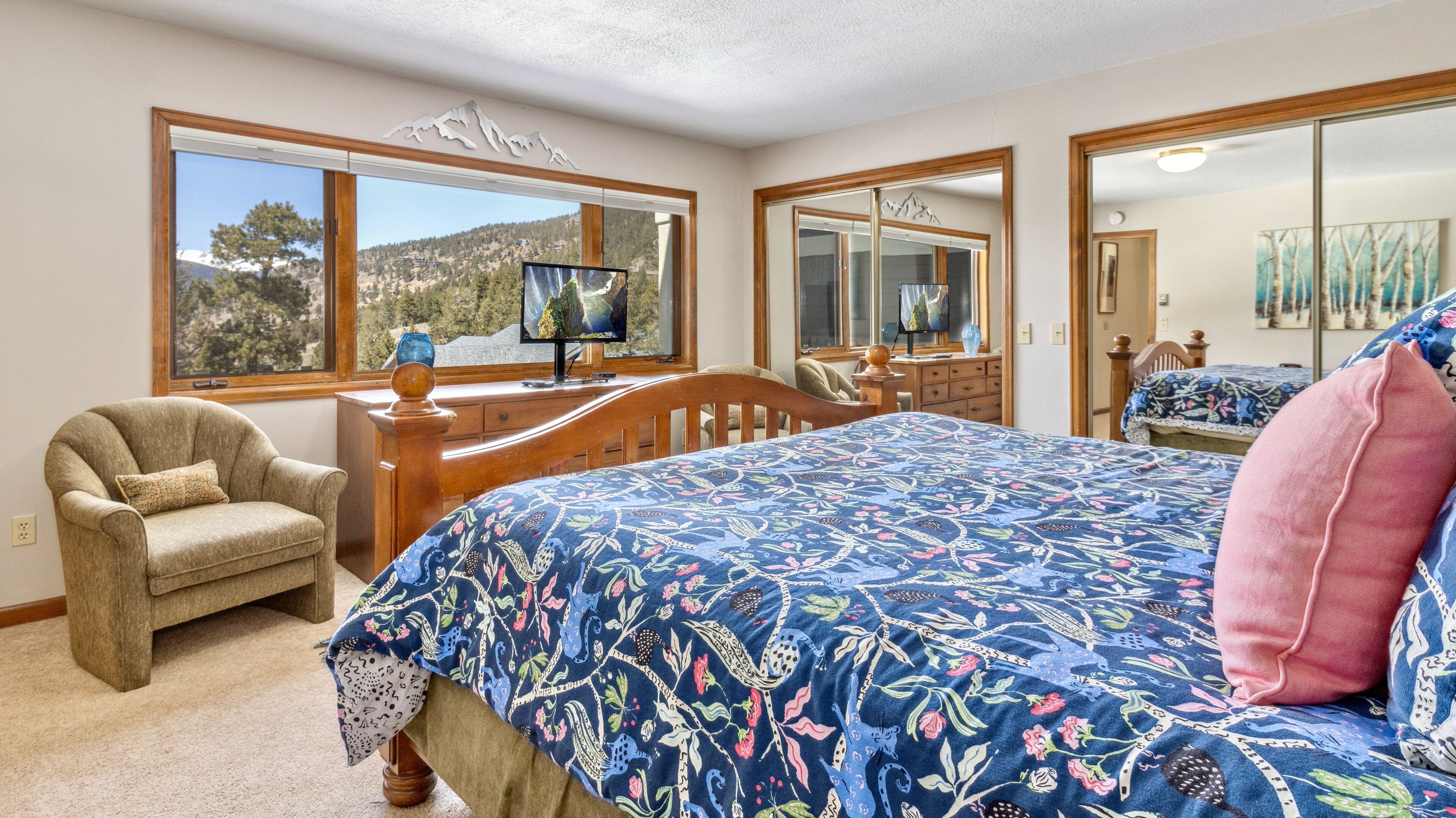 A bedroom with a large bed, a chair, a desk with a computer, and a large window with a mountain view.