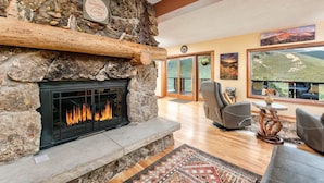 A stone fireplace with a fire, a stone shelf with decorations, a large window with a mountain view, and a cozy seating area with a rug. - Divine Destiny Luxury At Windcliff 6 Bedroom Condo by RedAwning (Estes Park)