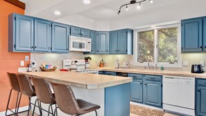 A kitchen with blue cabinets, a central island with bar stools, a microwave, a dishwasher, and a window with a view of trees. - Divine Destiny Luxury At Windcliff 6 Bedroom Condo by RedAwning (Estes Park)
