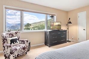 A bedroom with a large window, a bed, a dresser, a chair, and a lamp. - Wanderlust Luxury Vacation At Windcliff 3 Bedroom Condo by RedAwning (Estes Park)