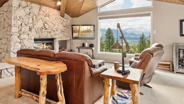 A cozy living room with a stone fireplace, leather sofa, and a large window offering a mountain view. - Wanderlust Luxury Vacation At Windcliff 3 Bedroom Condo by RedAwning (Estes Park)