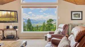 A cozy living room with a large window offering a mountain view, a leather sofa, a wooden coffee table, and a mounted TV. - Wanderlust Luxury Vacation At Windcliff 3 Bedroom Condo by RedAwning (Estes Park)
