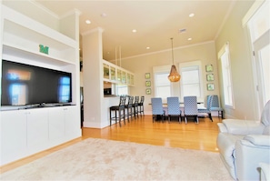 A modern living room with a flat-screen TV, a dining area with blue chairs, and a white sofa.