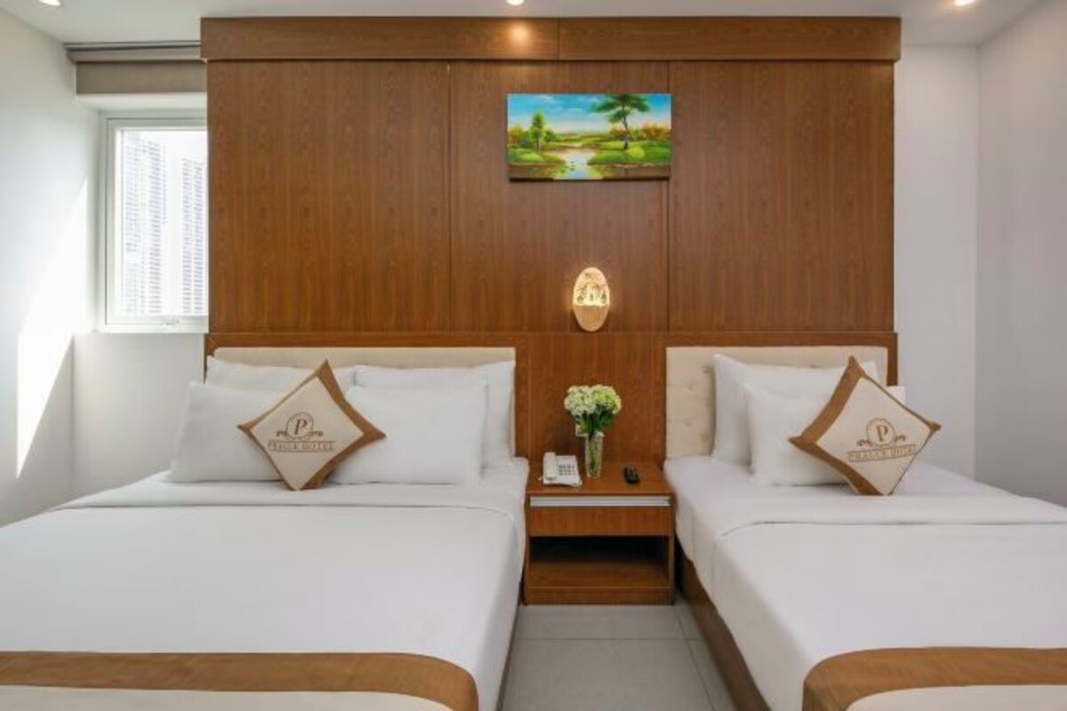 Quarto de hotel com duas camas, cabeceira de madeira, um quadro, mesa de cabeceira e um vaso com flores.