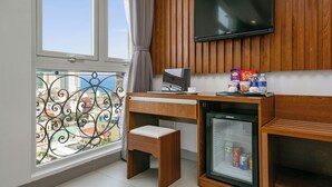 A compact room with a wooden desk, a small refrigerator, a TV mounted on the wall, and a window with a view of buildings. - Prague Hotel (Da Nang)